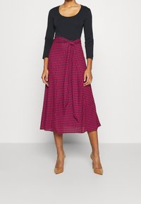 Svart långärmad topp kombinerad med en knälång rosa kjol med hounds-tooth mönster och en knytning vid midjan. Klassiska beige högklackade skor.