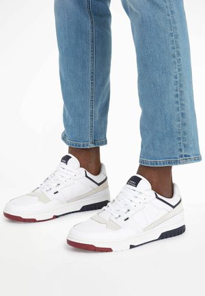 Białe sneakersy z teksturowanej skóry, kontrastowymi granatowymi akcentami i czerwoną gumową podeszwą. Nosi się je z niebieskimi dżinsami podwiniętymi na kostkach.