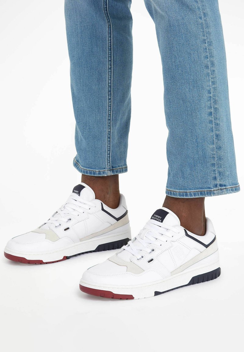 Białe sneakersy z teksturowanej skóry, kontrastowymi granatowymi akcentami i czerwoną gumową podeszwą. Nosi się je z niebieskimi dżinsami podwiniętymi na kostkach.