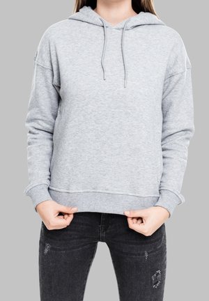 Personne portant un sweat à capuche gris clair et un jean noir déchiré, tenant l'ourlet inférieur du sweat à capuche avec les deux mains sur un fond uni.