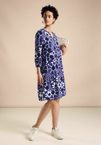 Blau-weißer Druck, Kleid mit Dreiviertelärmeln, V-Ausschnitt, gestuftem Rock und lockerer Passform, kombiniert mit hellen Sneakers und einer Clutch.