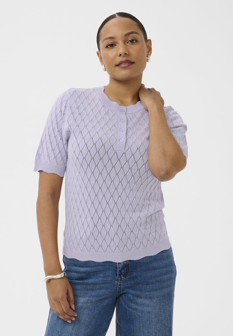 Femme portant un haut en maille lilas à manches courtes avec un motif en losange et trois boutons, associée à un jean bleu, posant avec la main sur le cou.