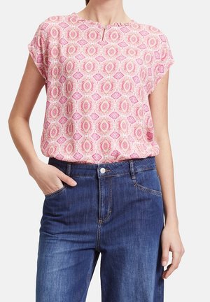 Kobieta w różowej bluzce z krótkim rękawem w wzory, włożonej do niebieskich dżinsów, z jedną ręką w kieszeni, a drugą spoczywającą przy boku.