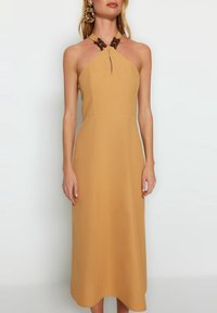 Ärmlös brun klänning med nyckelhålsurringning och distinkta mörkbruna flätade detaljer vid halsen. Slät tyg och midilängd.