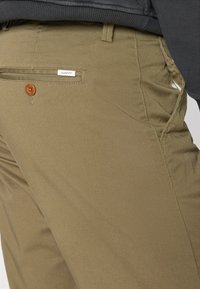 Olive green chinos made of cotton blend fabric feature a wooden button, side pockets, and a small brand label sewn on the back.