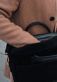 Black textured bag with a leather handle, partially unzipped, showing a green interior. A brown coat sleeve is visible holding the bag.