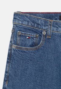 Blå denim jeans med bred midje, to frontlommer, kontrasterende oransje søm og metallnitter. Logopatch på den lille lommen.