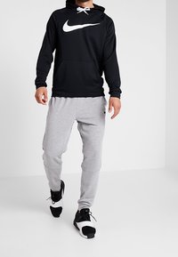 Uomo che indossa un hoodie nero Nike, pantaloni della tuta grigi chiari Nike e sneakers nero e bianche Nike mentre cammina in avanti su un pavimento bianco uniforme.