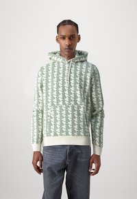 Felpa con cappuccio verde chiaro con un motivo geometrico, realizzata in tessuto morbido. Presenta una tasca frontale e polsini a costine. Indossata con jeans in denim scuro.