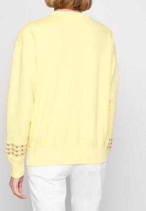 Personne portant un sweat-shirt jaune pâle avec une petite broderie florale aux poignets et un pantalon blanc, vue de dos.