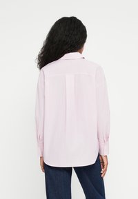 Chemise à manches longues de couleur rose clair avec un col à pointes, détail de pli dans le dos et poignets boutonnés, associée à un pantalon en denim foncé.