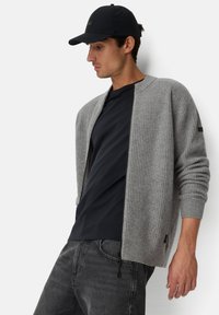 Grauer gerippter Cardigan mit Reißverschluss, getragen über einem marineblauen T-Shirt. Schwarze Mütze, graue Jeans und ein kleines Logo auf dem Ärmel.