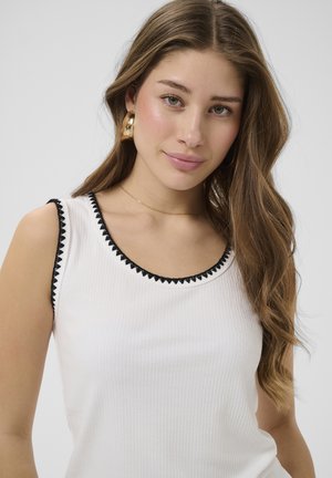 Tânără cu păr lung și castaniu, purtând un top alb fără mâneci cu margini negre și cercei mari rotunzi de aur, zâmbind ușor.