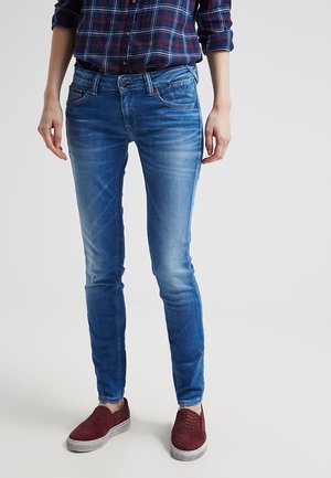 Persoon die een blauwe strakke spijkerbroek, een opgerolde ruitenoverhemd en rode instapschoenen draagt, staande op een lichte achtergrond.