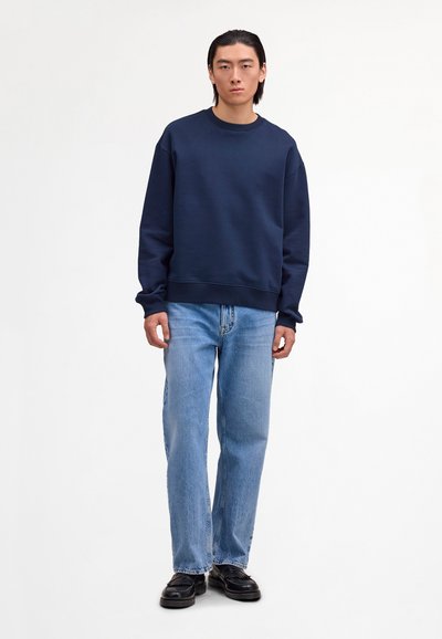 Young man standing wearing a navy crewneck sweatshirt, light blue straight-leg jeans, and black leather shoes on a white background.