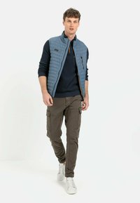 Hellblau gesteppte Weste über einem marineblauen Pullover, kombiniert mit olivgrünen Cargo-Hosen und weißen Sneakers. Mit Reißverschluss-Tasche und gerippten Bündchen.