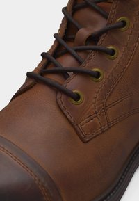 Κοντινό πλάνο καφέ δερμάτινης μπότας με μαύρα κορδόνια περασμένα μέσα από μπρούτζινα οπλισμένους κρίκους και λεπτομερείς ραφές κατά μήκος του άνω μέρους.