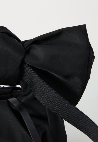 Bolsa de tela negra con un gran lazo en la parte superior, que presenta una superficie texturizada y una correa plana tejida para llevar.