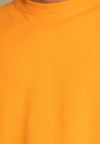 Oranssi puuvillainen t-paita, jossa on resorinen kaula-aukko ja kohokirjailtu 'P E C A D O R' -logo edessä. Sileä pinta ja normaali istuvuus.