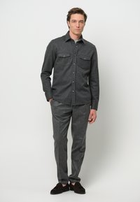 Camicia grigia con bottoni e due tasche sul petto, abbinata a pantaloni grigi coordinati e mocassini marrone scuro. Tessuto liscio e strutturato.