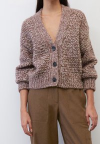 Růžový a krémový silný pletený kardigan s hlubokým véčkovým výstřihem, čtyřmi velkými černými knoflíky a širokými rukávy, nošený přes hnědé pouzdrové kalhoty.