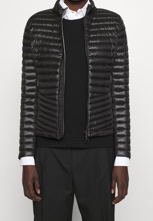 Man draagt een zwarte doorgestikte jas met rits over een zwarte trui en een wit overhemd met kraag, gecombineerd met een zwarte broek.