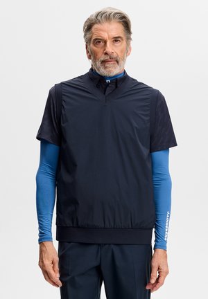 Reifer Mann mit grauem Haar und Bart, der blaue, geschichtete Sportkleidung trägt und vor einem schlichten weißen Hintergrund steht.