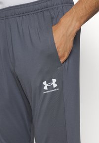 Ranka kišenėje tamsiai pilkų „Under Armour“ sportinių kelnių su elastingu juosmeniu ir matomu logotipu ant šlaunies.