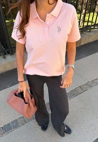 Femme portant un polo rose clair et un pantalon noir, tenant un sac à main mauve, debout sur le trottoir près d'une clôture en métal noire.