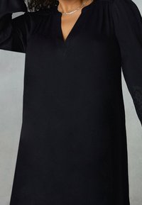 Černá tunika s výstřihem do V, dlouhé rukávy s krajkovým detailem na manžetách, vyrobená z hladké látky s volným střihem.
