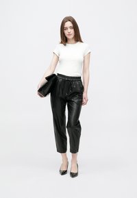 Naine, kes kannab valget lühikeste varrukatega toppi, mustast nahast pükse, musti kontsakingi, hoides suurt musta õlakotti valge ühevärvilise tausta ees.