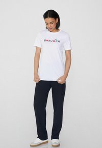 Bílý bavlněný tričko s potiskem "BONJOUR" v barevných písmenech, spárované s černými volnými kalhotami a bílými teniskami.