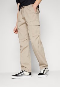 Béžové cargo kalhoty se bočními kapsami, elastickým pasem a šňůrkou na stažení. V kombinaci s černobílými teniskami se průhledným designem.