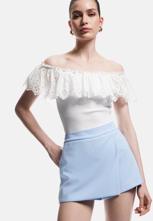 Mujer con blusa blanca de volantes sin hombros y falda mini envolvente de color azul claro, de pie con una mano en la cadera.