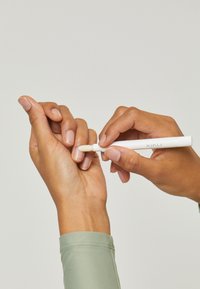Biały kosmetyczny długopis nakładany na dłoń. Długopis ma zwężoną końcówkę i jest trzymany przez palce z lekkim, naturalnym lakierem do paznokci.