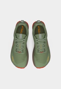 Ζευγάρι πράσινα παπούτσια για τρέξιμο στο μονοπάτι με γκρι κορδόνια, πορτοκαλί λεπτομέρειες και την επιγραφή "GORE-TEX" στην περιοχή των δακτύλων, φωτογραφημένα από πάνω.