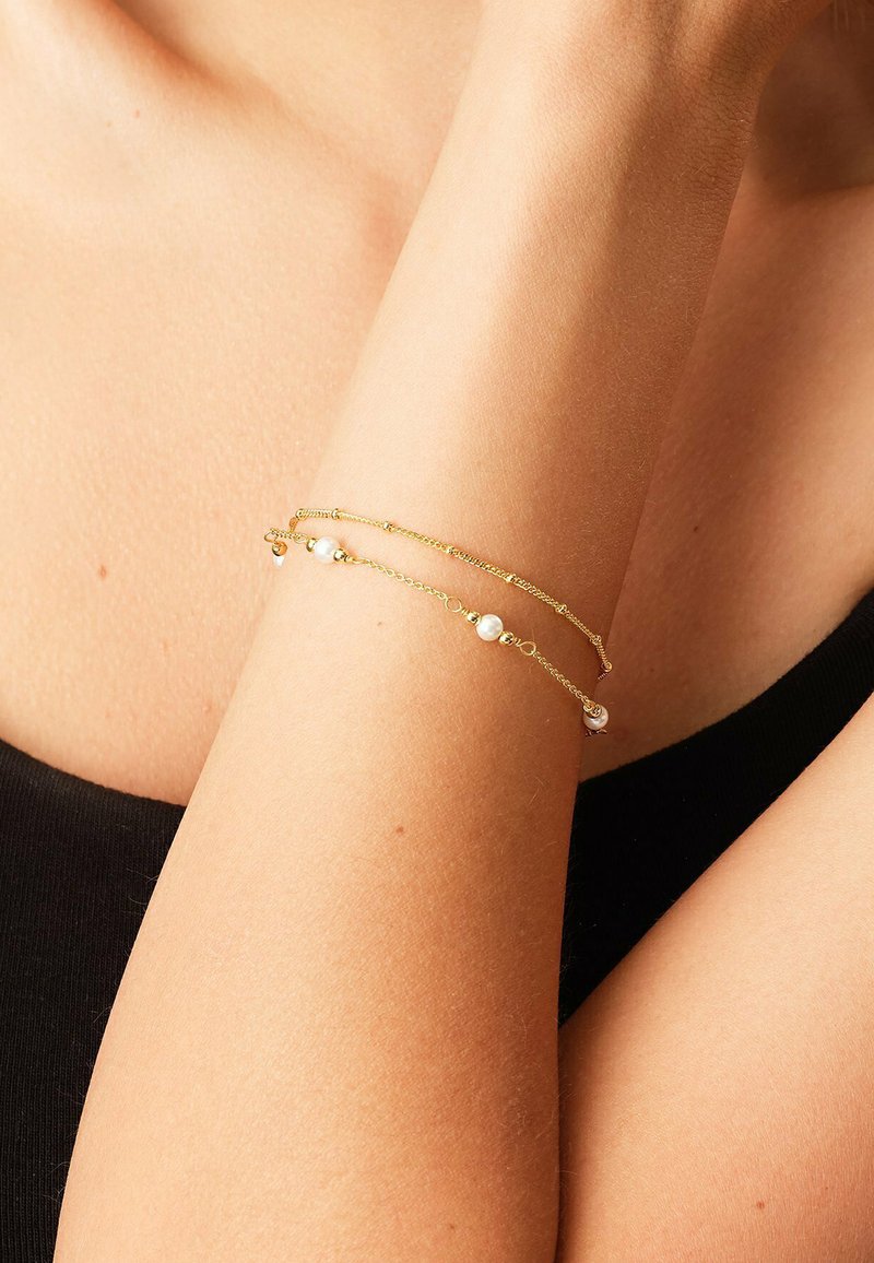 Tedere gouden armbanden met kleine parels dragen op een onderarm tegen huid en een zwarte stoffen achtergrond.