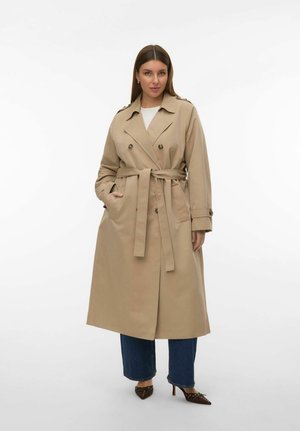 Kvinde iført en beige trenchcoat med bælte over en hvid top og blå jeans, stående med den ene hånd i lommen mod en ensfarvet baggrund.