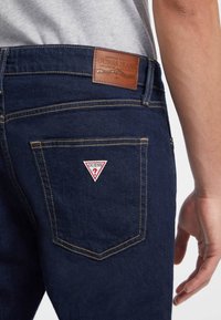 Tamno plave traperice s žutim šavovima, koje imaju kožnu Guess oznaku i crveni trokutasti logo na stražnjem džepu.