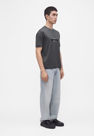 Homme debout portant un t-shirt gris foncé à manches courtes avec logo, un jean large bleu clair et des baskets noires sur un fond blanc uni.