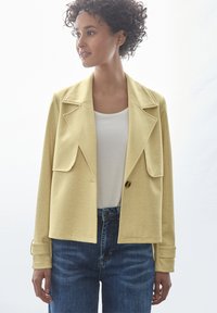 Blazer giallo a zigzag con grandi rever e due tasche frontali. Indossato su una maglietta bianca e abbinato a jeans di denim blu.