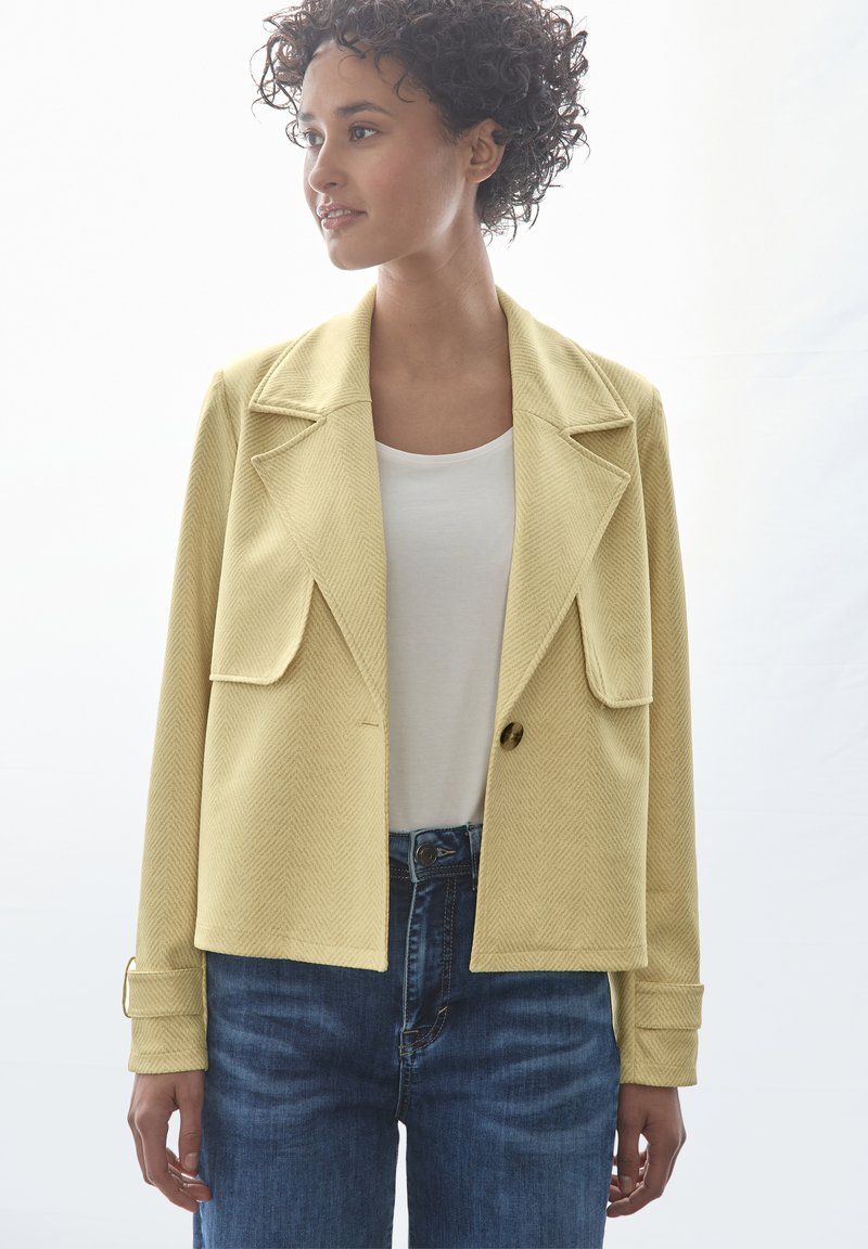 Blazer giallo a zigzag con grandi rever e due tasche frontali. Indossato su una maglietta bianca e abbinato a jeans di denim blu.