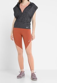 Mörkgrå huvlös ärmlös topp kombinerad med orange och persikofärgade leggings med geometriska paneldetaljer. Grå träningsskor fulländar looken.