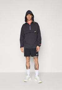 Schwarzer Pullover mit Kapuze, mit einem halben Reißverschluss vorne und mehrfarbigen Akzenten. Kombiniert mit schwarzen Shorts und weißen Sneakern.