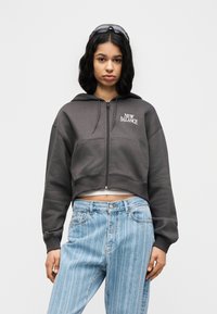 Νεαρή γυναίκα που φορεί φούτερ με φερμουάρ σε σκούρο γκρι με το λογότυπο «New Balance» και ανοιχτόχρωμο τζιν με ρίγες, στέκεται μπροστά σε απλό λευκό φόντο.