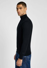 Maglione nero a maglia con colletto alto, maniche lunghe e orli a coste. Il tessuto sembra liscio e leggermente aderente. Abbinato a jeans in denim blu.