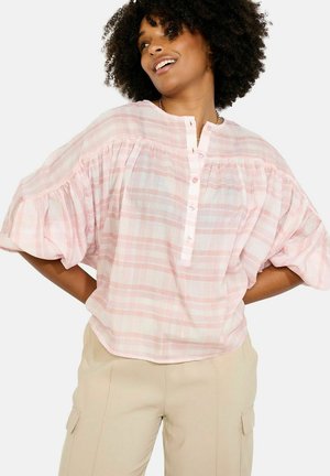 Žena nosi priesvitnú ružovo-bielu károvanú blúzku s gombíkmi a béžové nohavice, usmieva sa s rukami v bok na čistom pozadí.