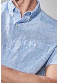 Chemise en lin bleu clair à manches courtes avec col boutonné, dotée de deux poches poitrine et d'une texture subtile sur l'ensemble du tissu.