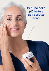 Un contenitore rotondo di crema bianca etichettato "NIVEA VITAL", tenuto da una persona che sta applicando la crema sul viso con una carnagione liscia e chiara.