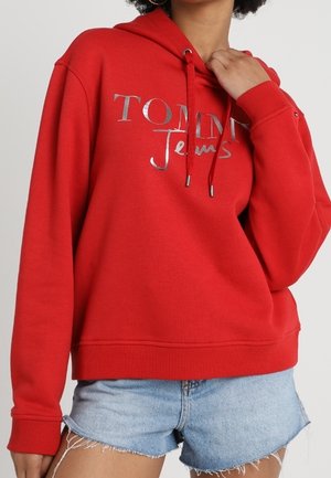 Kvinde iført rød Tommy Jeans hoodie og lyseblå denimshorts, der holder i hoodien nær halsudskæringen.